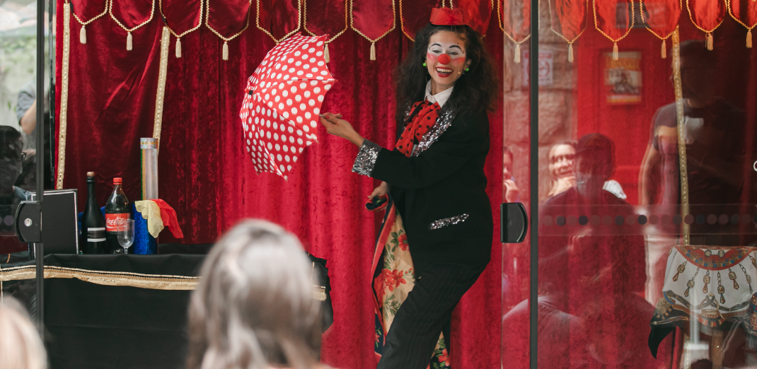 Na imagem está uma mulher vestida de palhaça sorrindo e segurando um guarda-chuva vermelho de bolinhas enquanto performa para o dia das crianças.