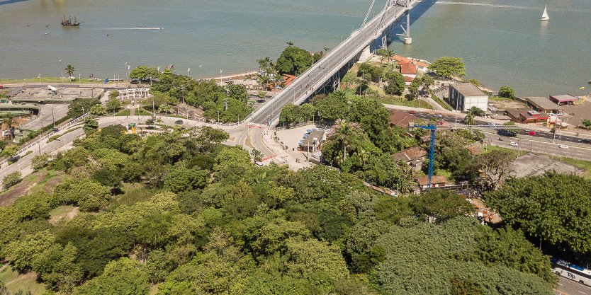 A imagem apresenta uma perspectiva aérea do Parque da Luz em Florianópolis, destacando o encontro da natureza com a urbanização. A ponte, um ponto focal da cena, atravessa tranquilamente a água, conectando as áreas verdes do parque com o tecido urbano adjacente.