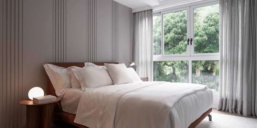 Showroom: Quarto de suíte com cama de casal, lençóis brancos e cabeceira de madeira. Janela ampla com vista para área verde, acompanhada de cortinas cinza, proporcionando privacidade e controle de luz natural.