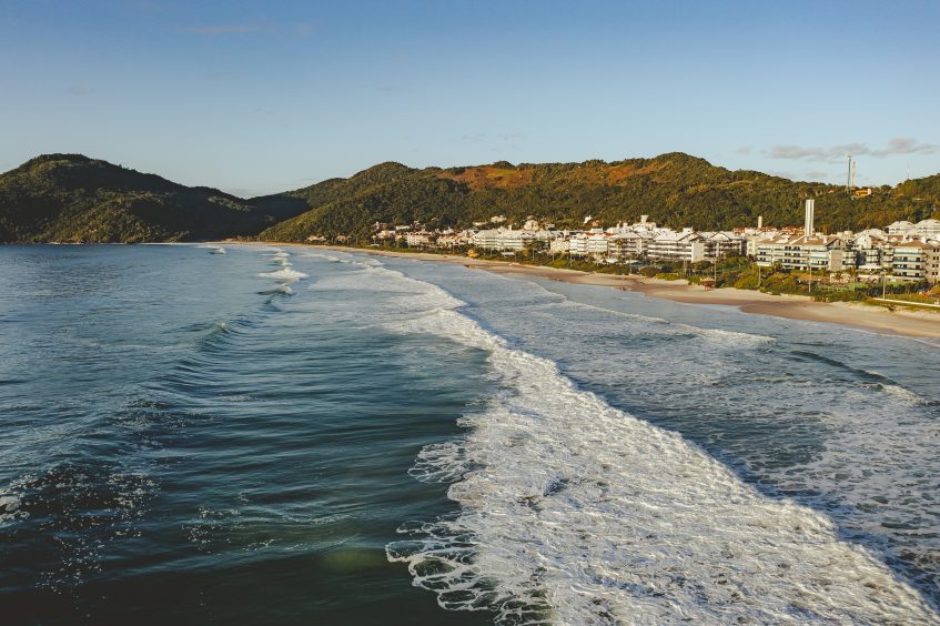 A foto mostra o registro de uma praia em Florianópolis, onde podemos ver o mar e a areia, ao fundo vemos alguns empreendimentos.